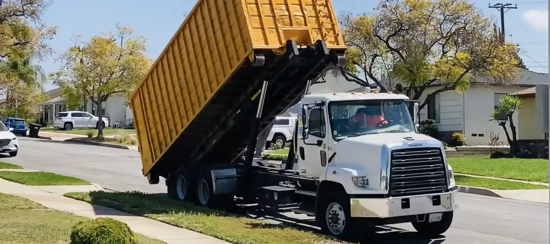 A roll-off truck tipping a large yellow dumpster into place on a residential street.