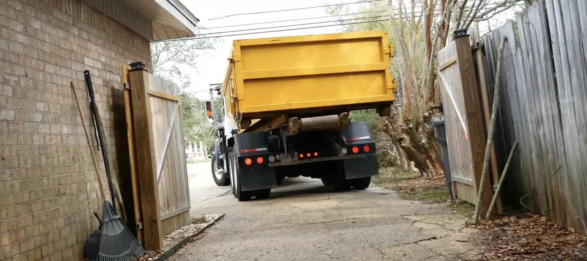 A roll-off delivery truck backing a dumpster through a narrow gated alley to reach a home.