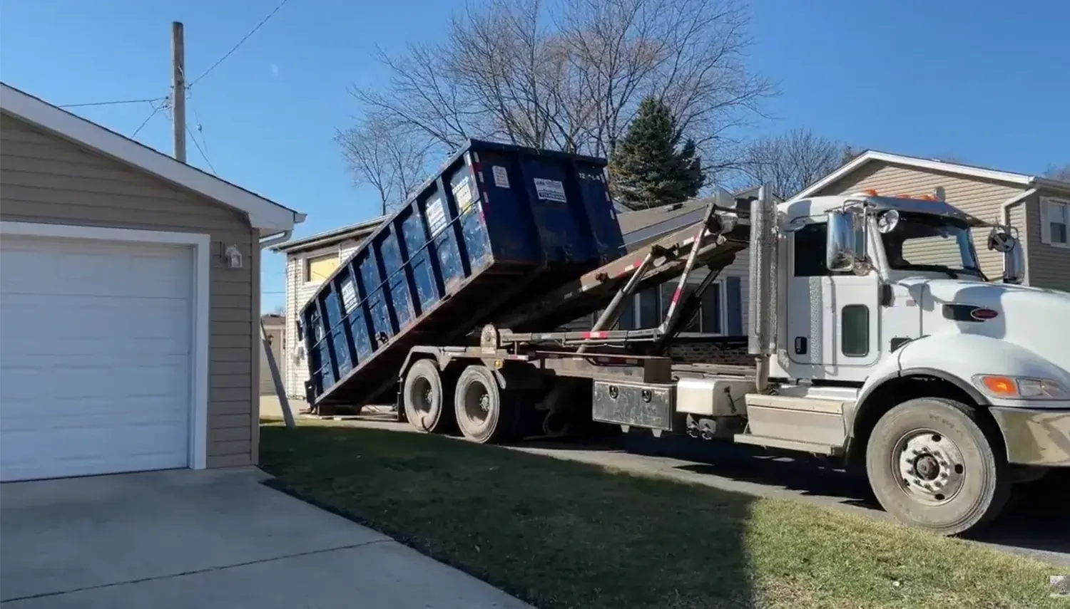 A roll-off dumpster truck backing into a residential driveway for delivery