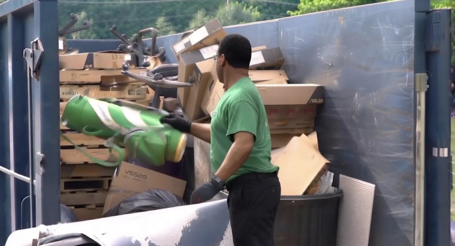 A worker loading office furniture and boxes into a commercial roll-off dumpster.