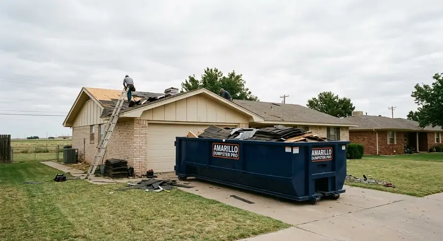 A roll-off dumpster on a driveway during a roof tear-off, with a crew removing asphalt shingles.